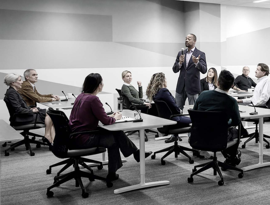Man giving a presentation in a conference room filled with guests that have wireless microphones in front of them.