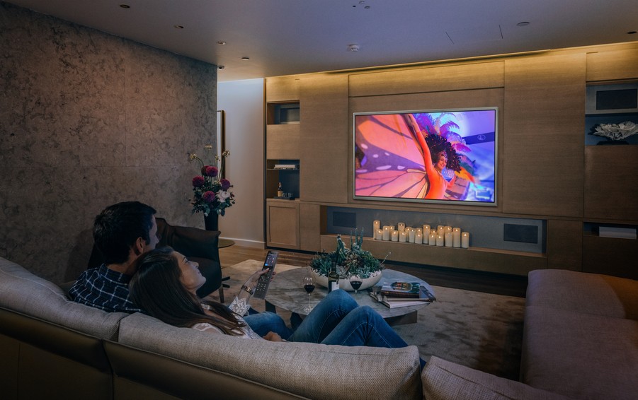 Couple sits on a couch in a dimmed home theater, looking at large TV mounted on the wall.
